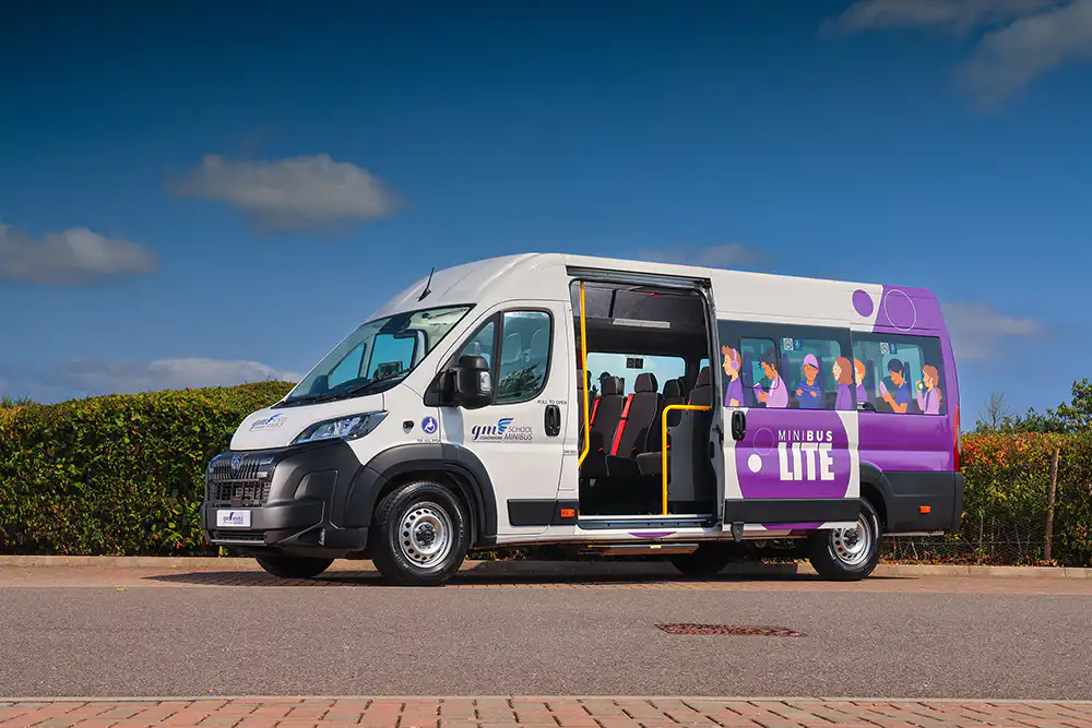 Side angle view of a white GM Coachwork Minibus Lite with purple graphics and cartoon student illustrations, parked outdoors with its side passenger door wide open to reveal interior seating.