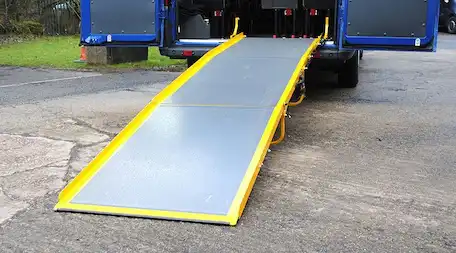 Rear view of an open blue wheelchair-accessible van with a long grey access ramp extended to the ground, bordered by bright yellow safety side rails.